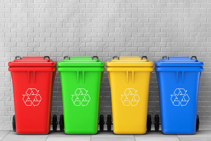 Four recycling containers, in red, green, yellow, and blue, in a row against a gray brick wall.