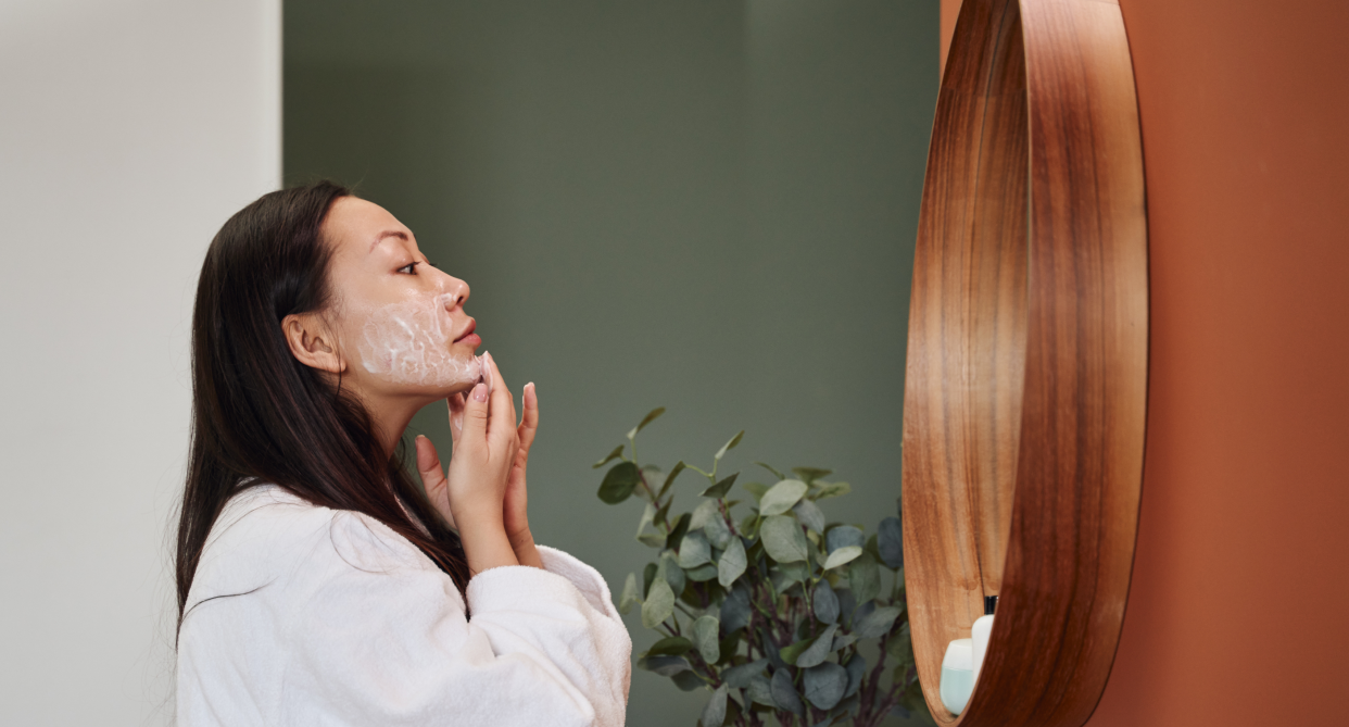 asian woman with long black hair wearing white bath robe and face retinol anti-aging cream looking in mirror