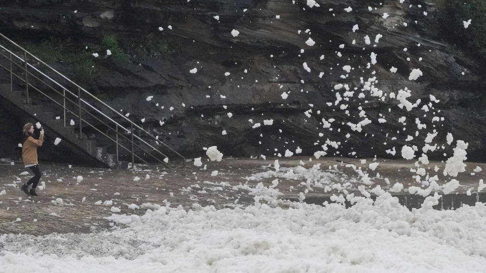 A woman films as sea foam is blown onto her at Bronte Beach as rain continues to fall in Sydney, Australia, Wednesday, July 6, 2022. More than 50,000 residents of Sydney and its surrounds have been told to evacuate or prepare to abandon their homes on Tuesday as Australia's largest city braces for what could be its worst flooding in 18 months. (AP Photo/Mark Baker)