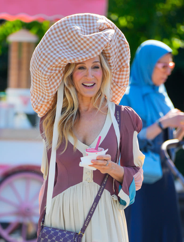 Sarah Jessica Parker wearing a large checkered hat, a stylish long-sleeved dress, and holding a cup of frozen yogurt. Another person is in the background