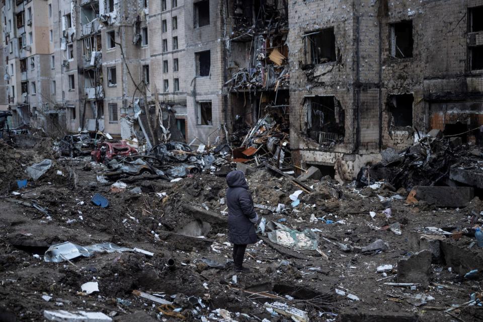 A woman in a dark coat stands on torn-up ground and looks up a heavily damaged multistory building