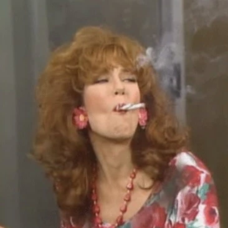 A woman with curly hair wearing a floral-patterned top, red necklace, and large earrings smokes a cigarette, blowing smoke up and to the side