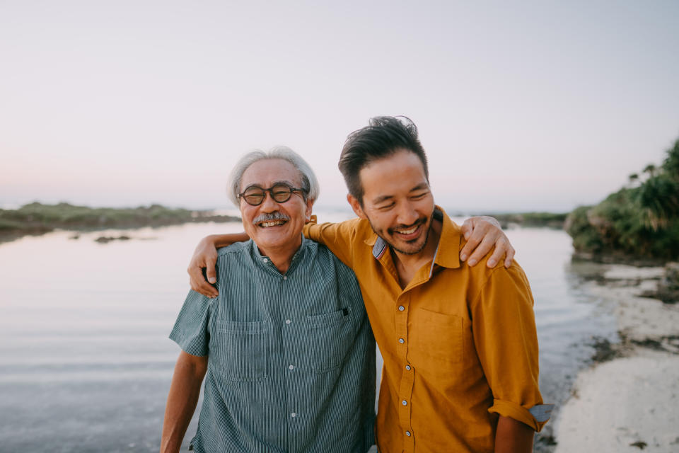 Two people, an elderly man, and a younger man, smile while standing by a body of water with arms around each other. The older man wears glasses and a short-sleeve shirt, and the younger man wears a long-sleeve shirt