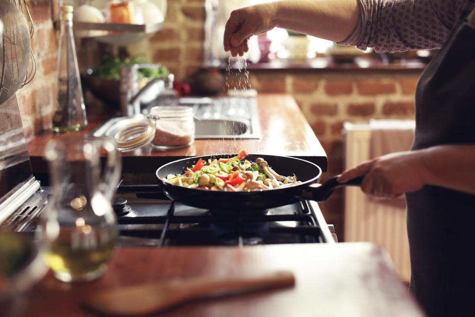 A person sprinkles seasoning into a skillet filled with vegetables, cooking on a stove in a rustic kitchen