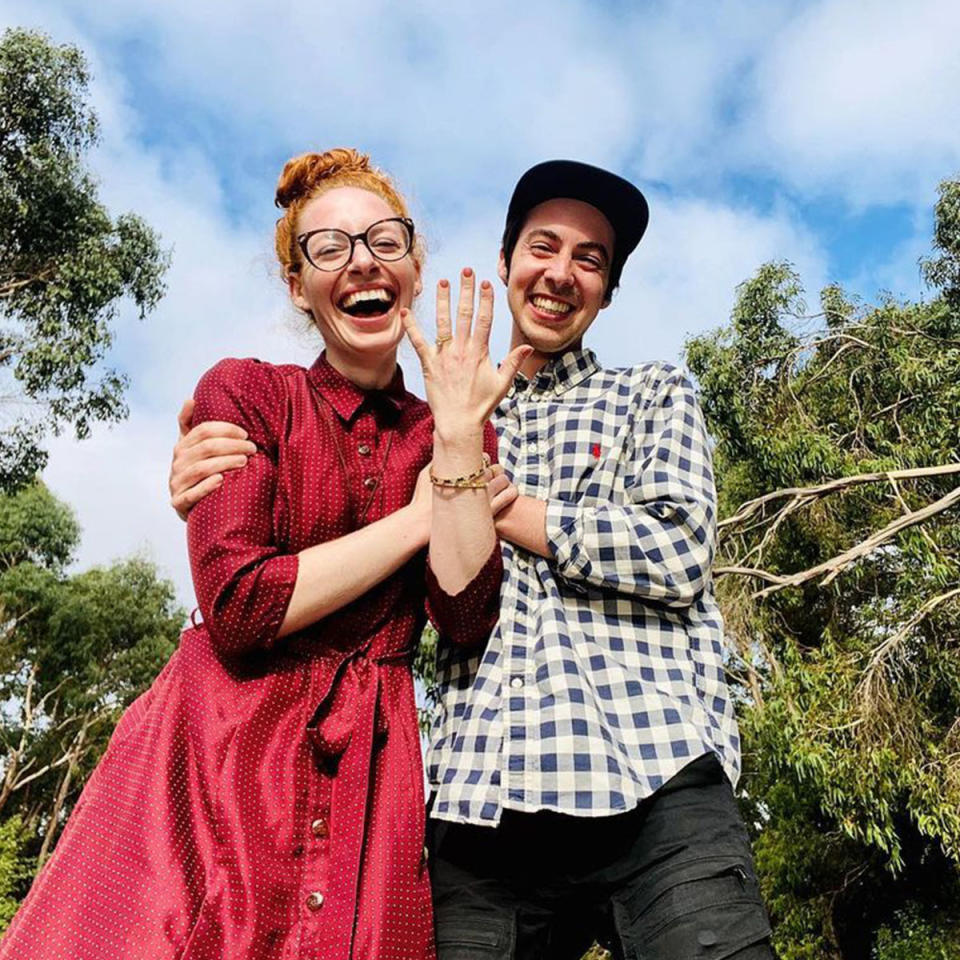 Emma Watkins in a red dress, glasses, and holding up her hand to show off her engagement ring, next to Oliver Brian in a checkered shirt and black hat.