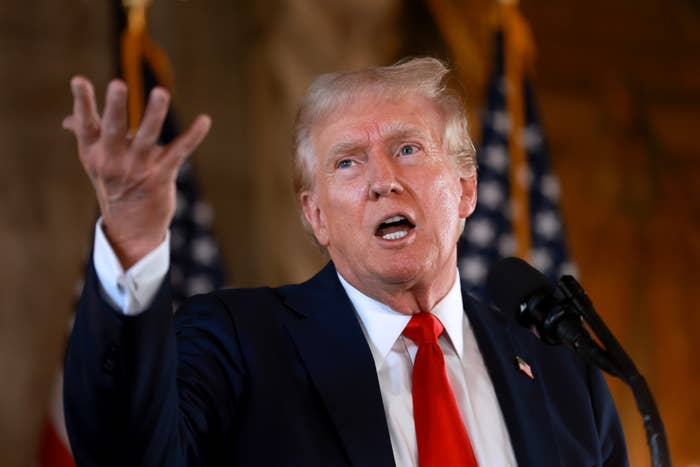 Donald Trump speaking passionately at a podium in front of U.S. flags, wearing a dark suit, white shirt, and red tie