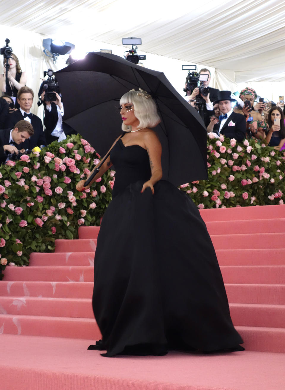 Lady Gaga stands on a pink staircase at a formal event, wearing a voluminous black gown and holding a black umbrella. Photographers are in the background