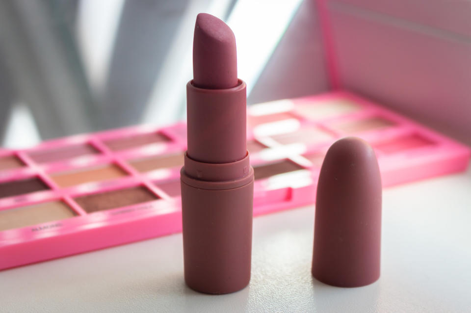 An open tube of lipstick with the cap sitting next to it and an eyeshadow palette in the background