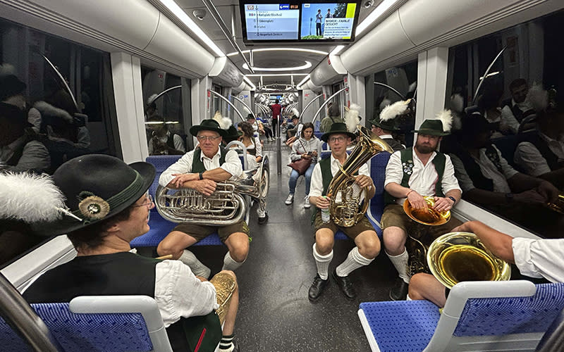 Bavarian musicians sit in a subway on their way to a traditional costume. The photo is taken from an angle looking down the aisle at musicians seated to the left and right. Some of them hold large brass instruments on their laps or on the floor between their legs. The musicians are wearing traditional German costumes.