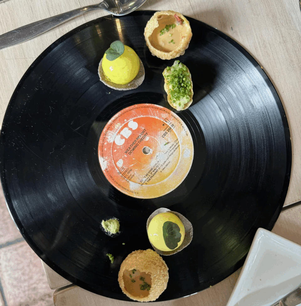 A black vinyl record is used as a serving plate for several small, artistic food items, including round yellow spheres and a crispy shell with green garnish