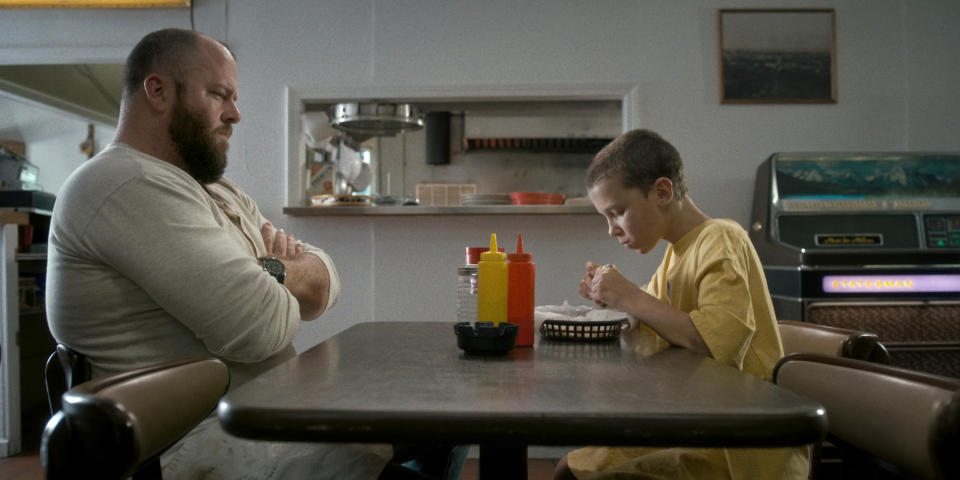 a grown man looking grumpy watches and sits across a small kid eating in a dinner