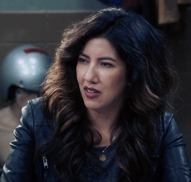 Stephanie Beatriz, dressed in a black leather jacket, appears in a scene from the TV show Brooklyn 99. She has long wavy hair and wears a pendant necklace