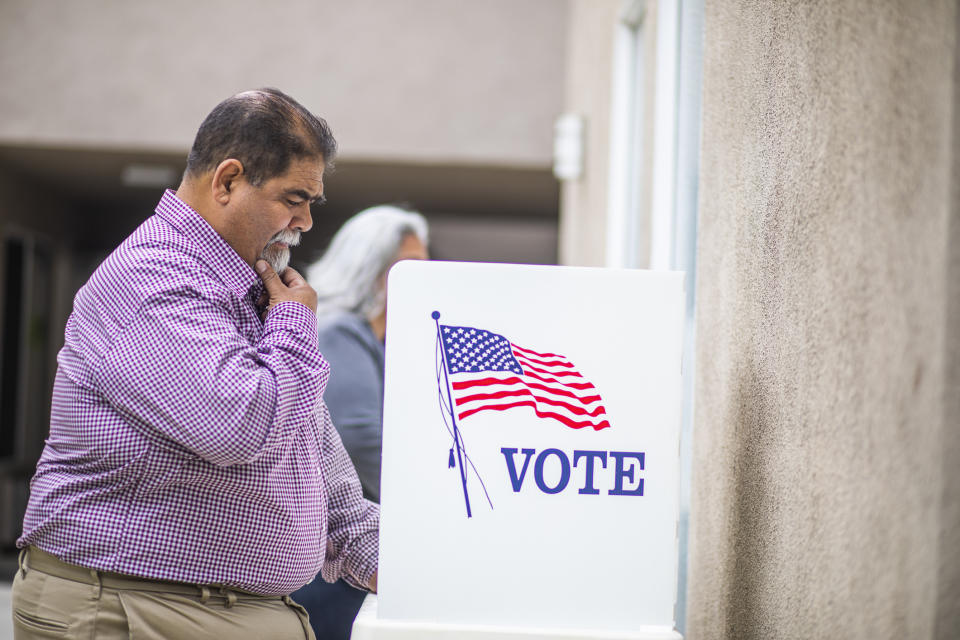 A man in a checkered shirt stands thoughtfully at a voting booth with an American flag and the word "VOTE" on it. Another person is partially visible behind him