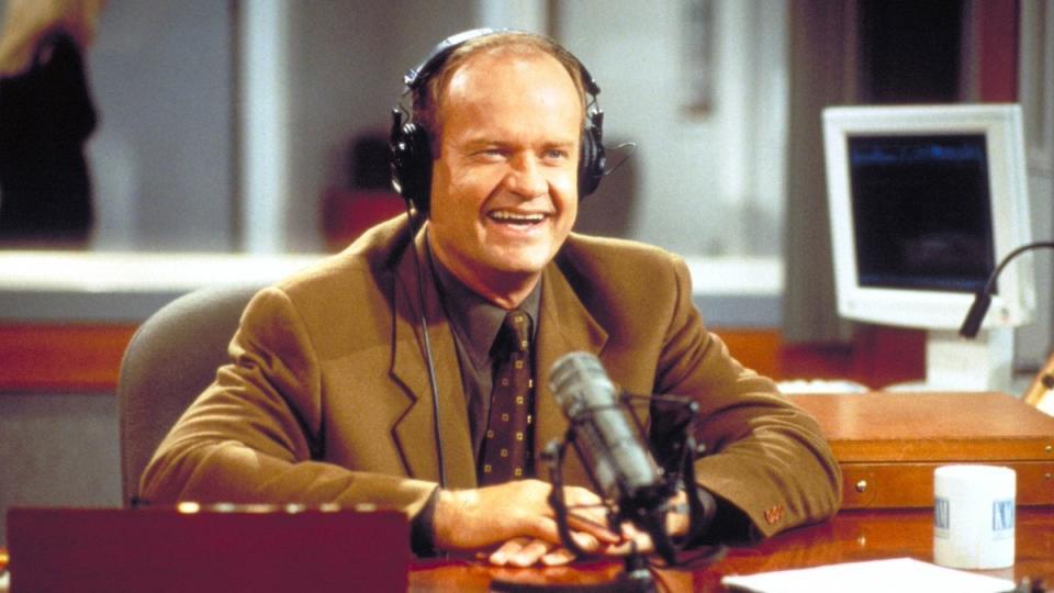 A still of Frasier Crane from the TV show, he's in a recording studio wearing headphones and smiling with a mic in front of him.