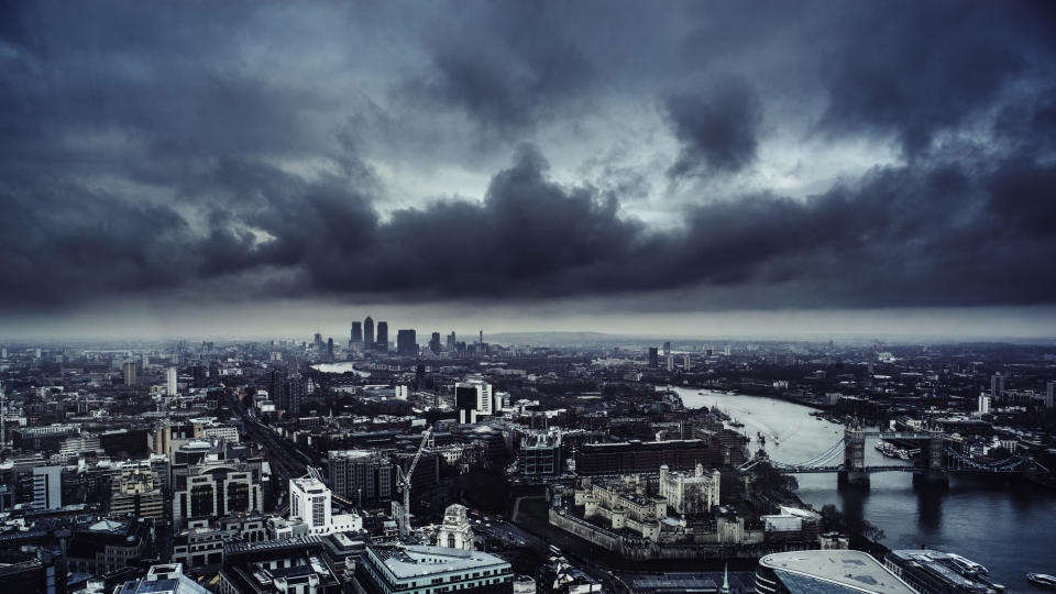 Aerial view of London looking down the River Thames towards Canary Wharf and the Docklands. In the foreground is the Tower of London and Tower Bridge. A dark and menacing sky is overhead.