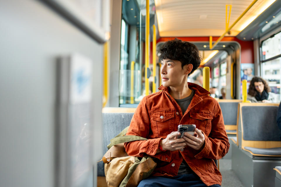 A person sits on a train holding a phone, gazing out the window. They are wearing a casual jacket. The background includes other passengers and the train's interior