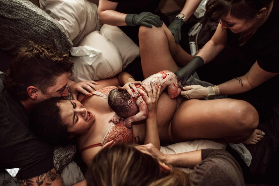 Newborn rests on mother's chest immediately after birth, with father embracing them and medical staff assisting