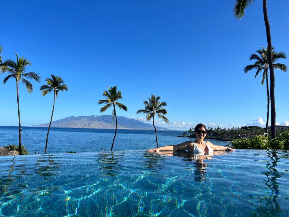 A woman poses in a pool with the ocean and mountains behind her.
