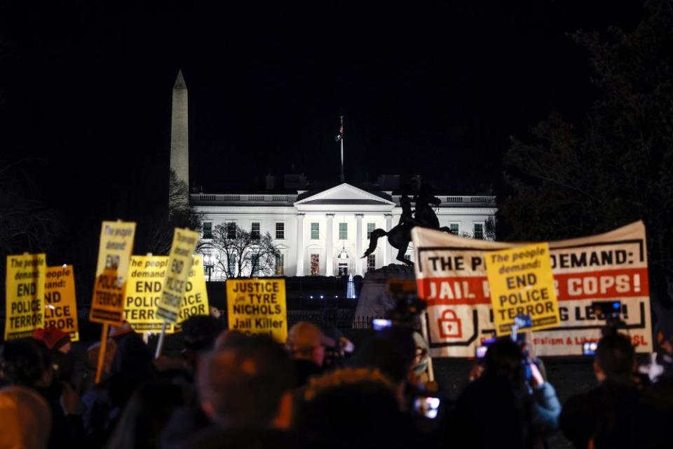 People take part in an evening protest holding signs reading ‘End police terror’ with the White House lit up in the background.