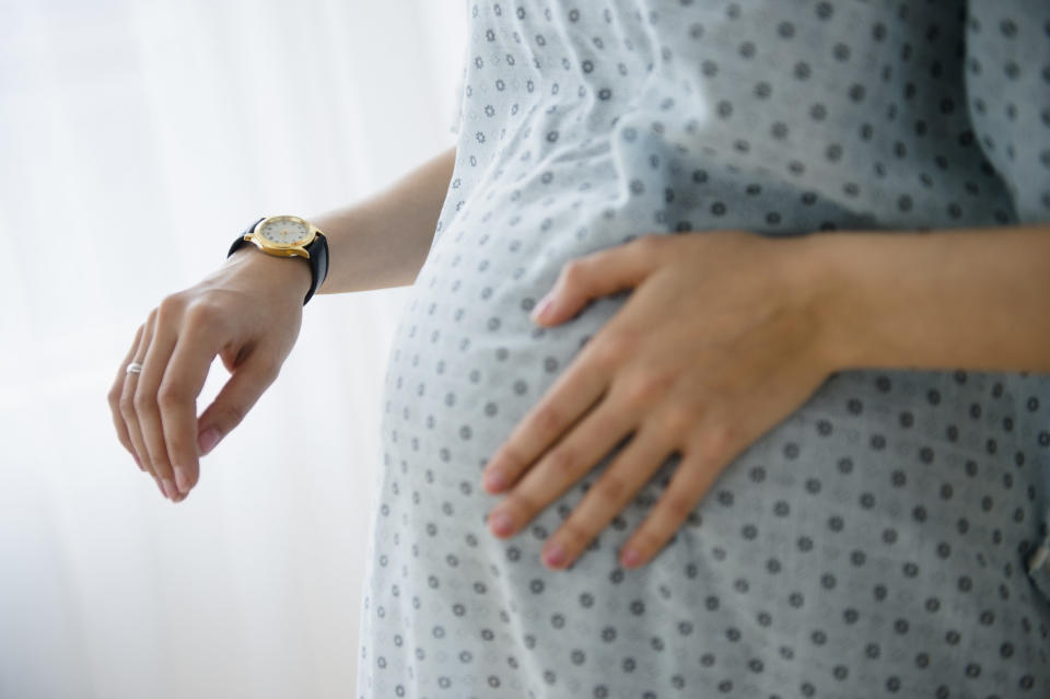 A pregnant woman holds her stomach and looks at a watch while wearing a hospital gown