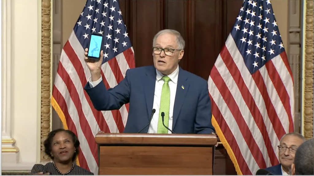 Washington Gov. Jay Inslee displays a photo of the Columbia River as White House Council on Environmental Quality Chair Brenda Mallory and White House senior advisor John Podesta look on before a Feb. 23, 2024, White House signing ceremony for an agreement to protect the river. (Screenshot from White House live feed.)