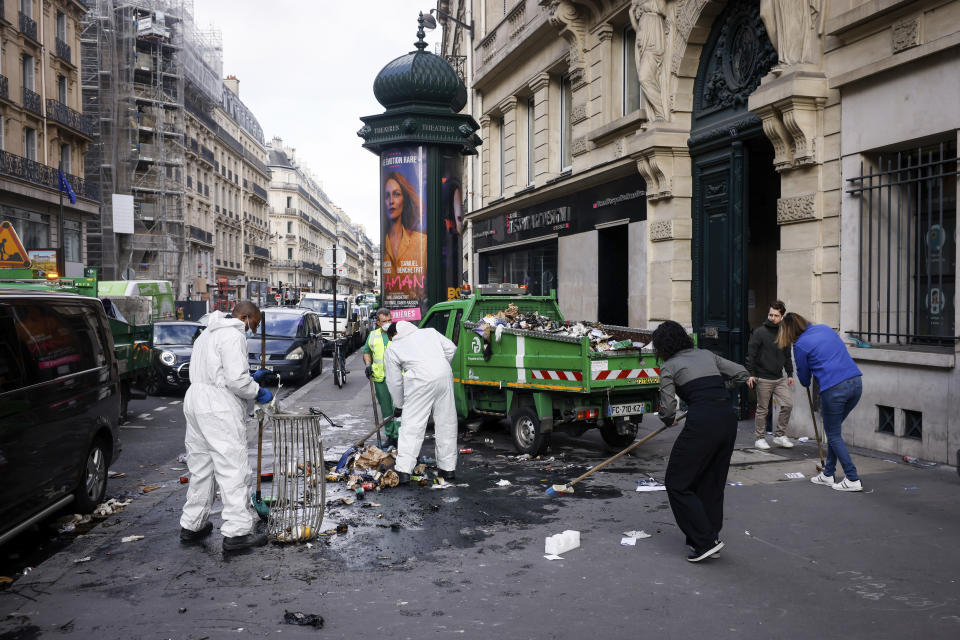 Municipality workers and two women cleaning a street from burned garbage of last night protests against the retirement bill in Paris, Friday, March 24, 2023. French President Macron's office says state visit by Britain's King Charles III is postponed amid mass strikes and protests. (AP Photo/Thomas Padilla)