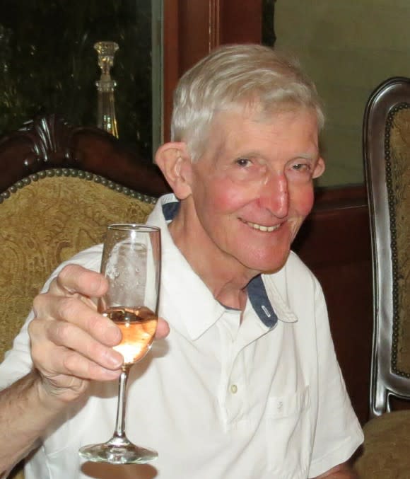 Man (Hayford Peirce) smiling at camera, toasting with a champagne glass. He is wearing a white polo with a white undershirt, sitting in a chair at a table, in front of what appears to be a candle holder in a case.