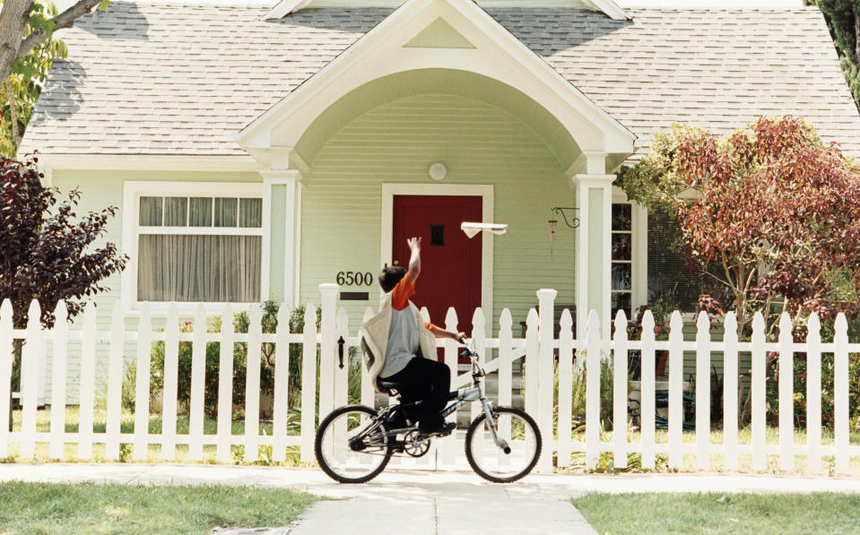 A young person riding a bicycle is tossing a newspaper in front of a house