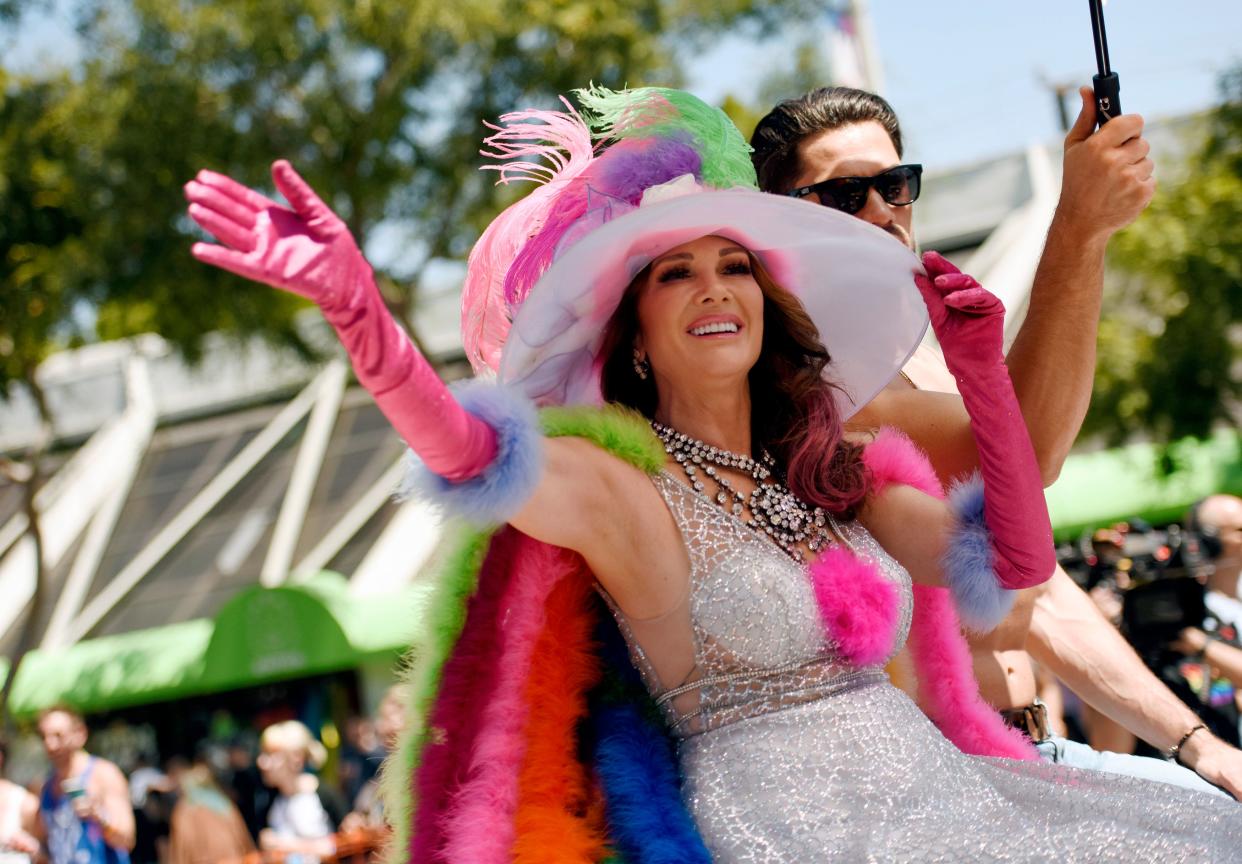 Lisa Vanderpump sits on a parade float waving and holding onto her colorful hat