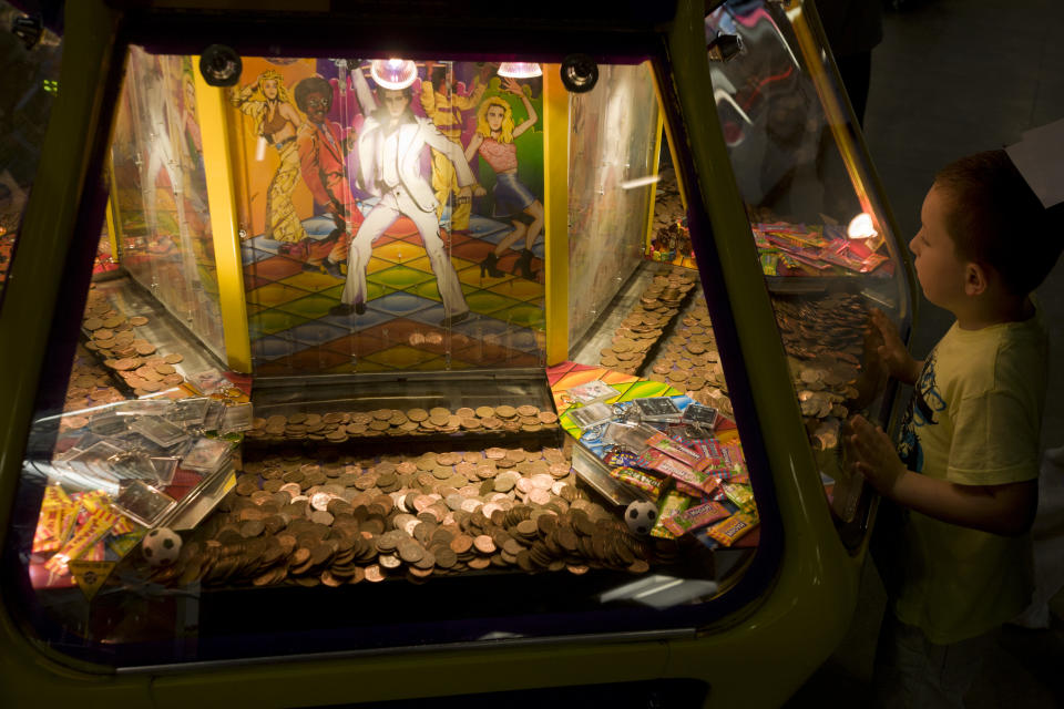 A young boy watches the sweeping arms of a Penny push/fall game in the amusement arcade at Weston-super-Mare's grand pier. Waiting for some coins to be caught in the log-jam and to fall into the prize tray below, the lad seems spellbound by the potential luck and possibilities although the odds are against him. Images of 1970s dancers, including John Travolta, strut their stuff at the disco. 2p pennies stack up until the moment when they topple over and spill out. (Photo by In Pictures Ltd./Corbis via Getty Images)