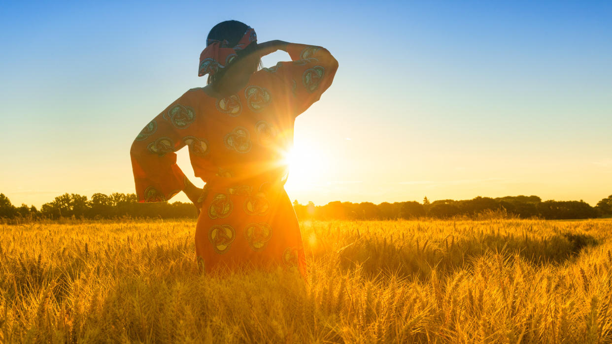 African woman in traditional clothes standing, looking, hand to eyes, in field of barley or wheat crops at sunset or sunrise.