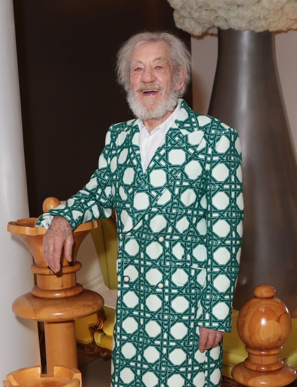 ian mckellen smiles as he stands in a hotel lobby with his arm leaning on a giant chess piece