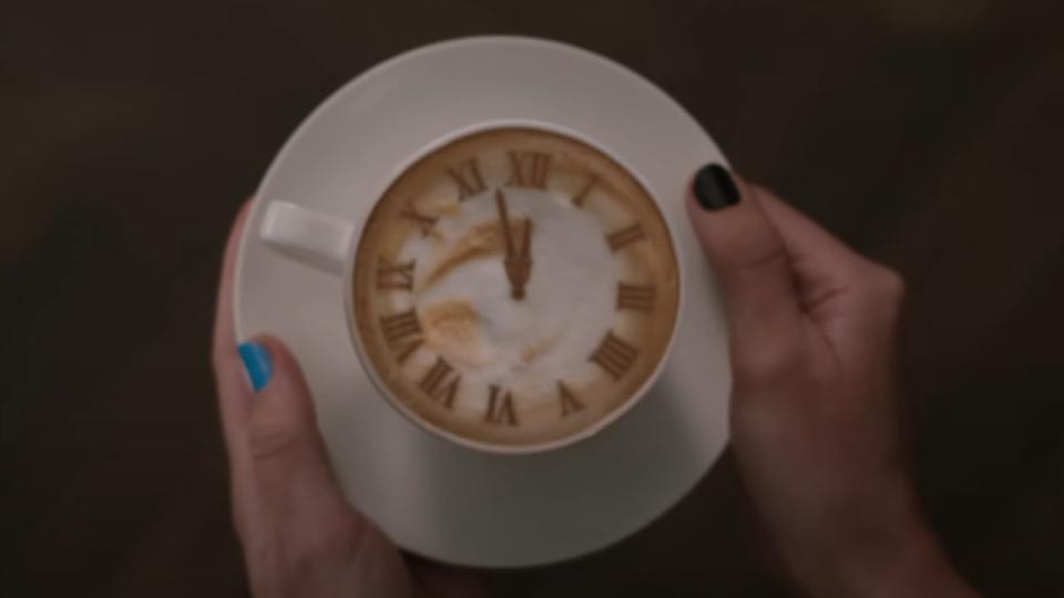 Taylor Swift's hands with blue and black nails holding a coffee cup with latte art of a clock that is about to strike midnight.