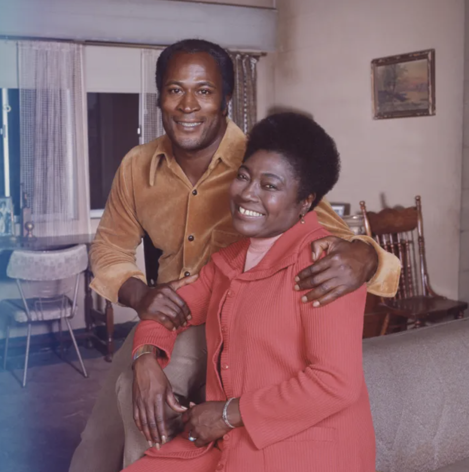 Two people smiling, seated closely on a couch, both dressed in buttoned shirts