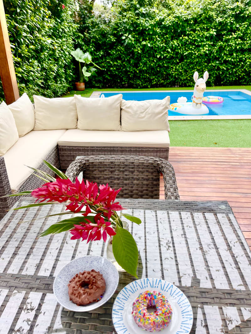 Doughnuts and flowers on glass top woven table and chair on deck overlooking pool.