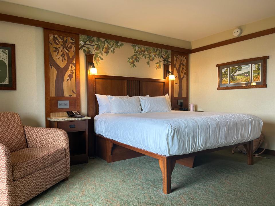 A hotel room with a king bed with white linens, a patterned chair, two nightstands with pear-tree murals, and a mural of a window.