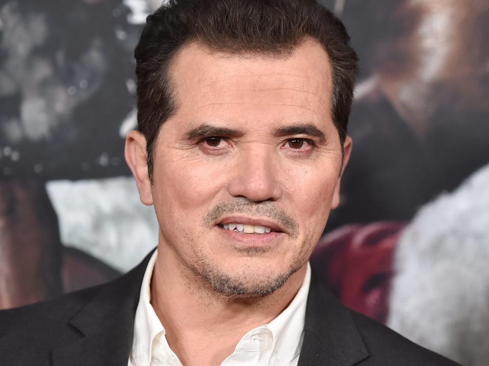 John Leguizamo poses for photos in a black suit jacket and white shirt.