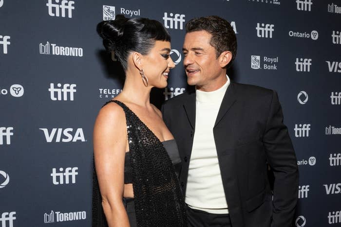 Katy Perry in a black sequined dress and Orlando Bloom in a black suit smile at each other on the red carpet at TIFF