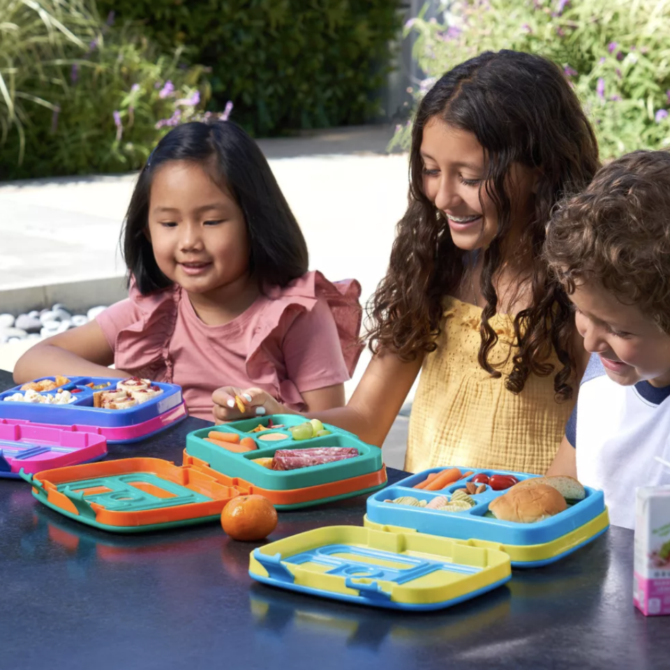 Children enjoying a meal from colorful bento lunch boxes; one girl wears a pink dress, another wears a yellow top, and a boy wears a blue and white striped shirt