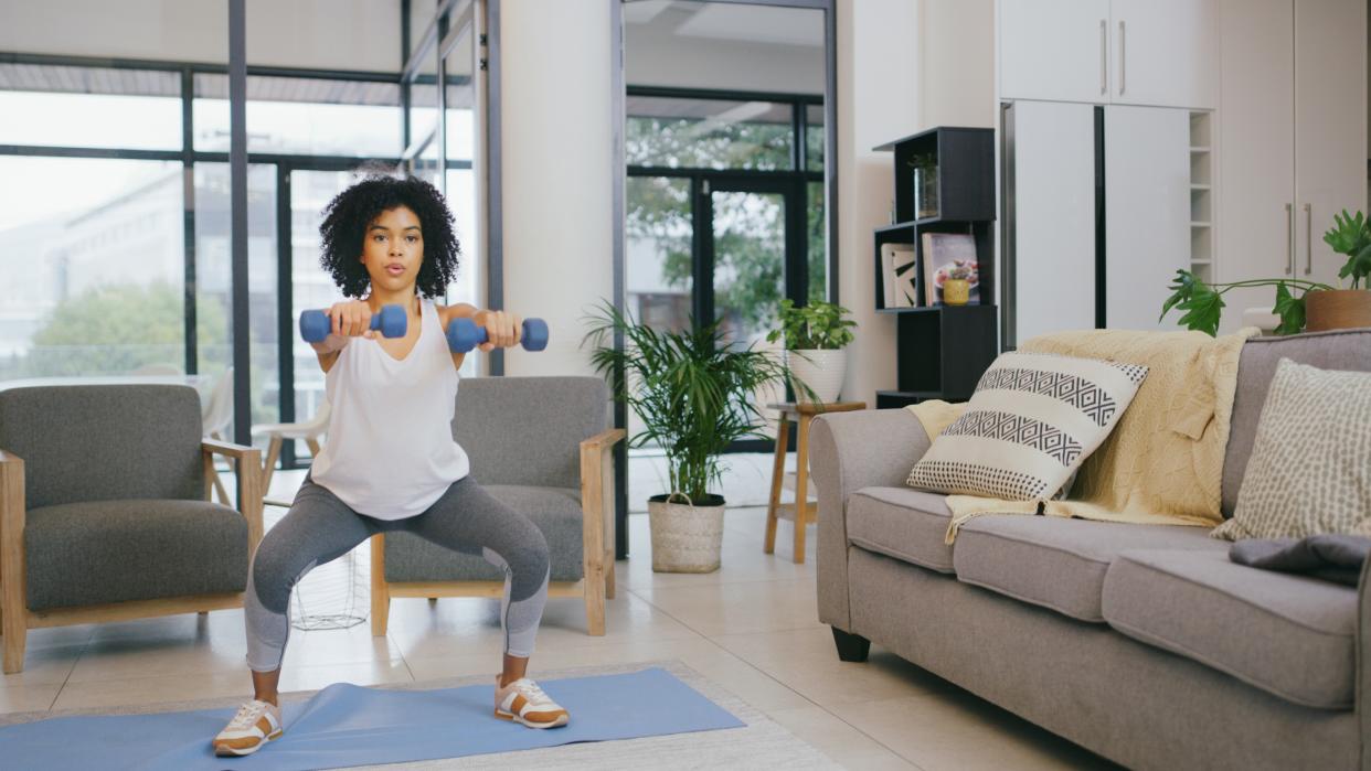  Woman in workout clothes doing a squat with two dumbbells in a stylish living room. 