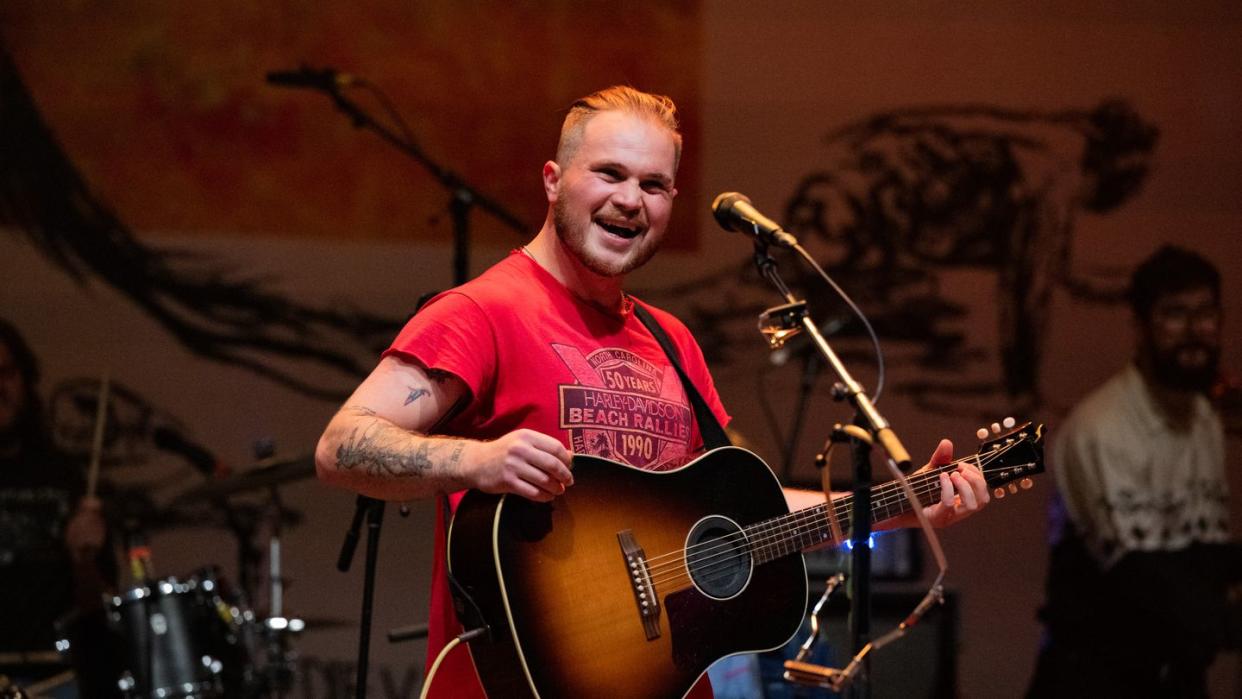 zach bryan smiling behind a microphone while playing his guitar