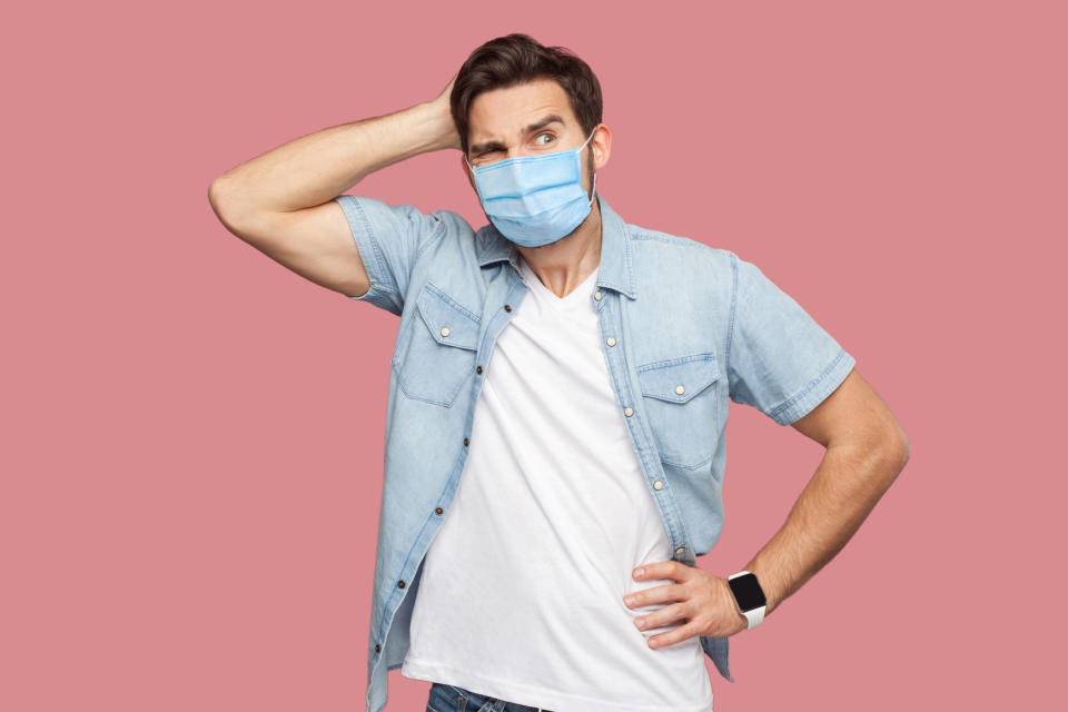 Portrait of young man with surgical medical mask in blue casual style shirt standing, scratching his head, looking away and thinking what to do.