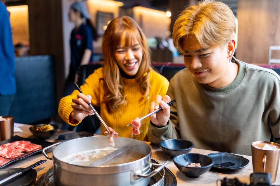A man and a woman are enjoying a hot pot meal together at a restaurant, smiling and using chopsticks to cook ingredients in a central pot
