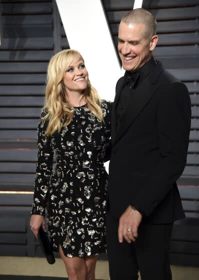 Reese Witherspoon in a black dress looking to a man on her right wearing a black suit