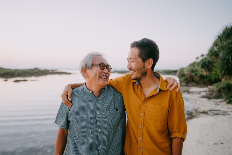 Two smiling men, potentially father and son, standing close outdoors with water and sky in the background