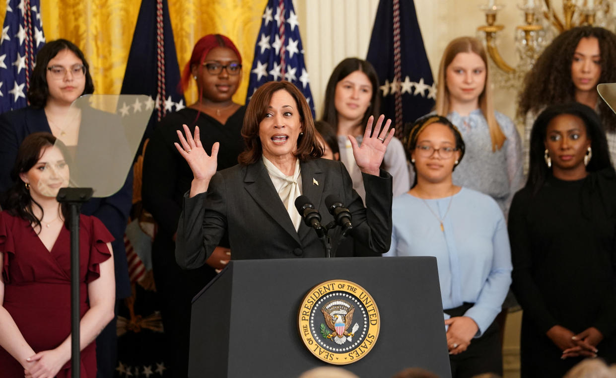 Vice President Kamala Harris at a podium marked Seal of the President of the United States, raising both hands in surprise, with two rows of young women arrayed behind her.