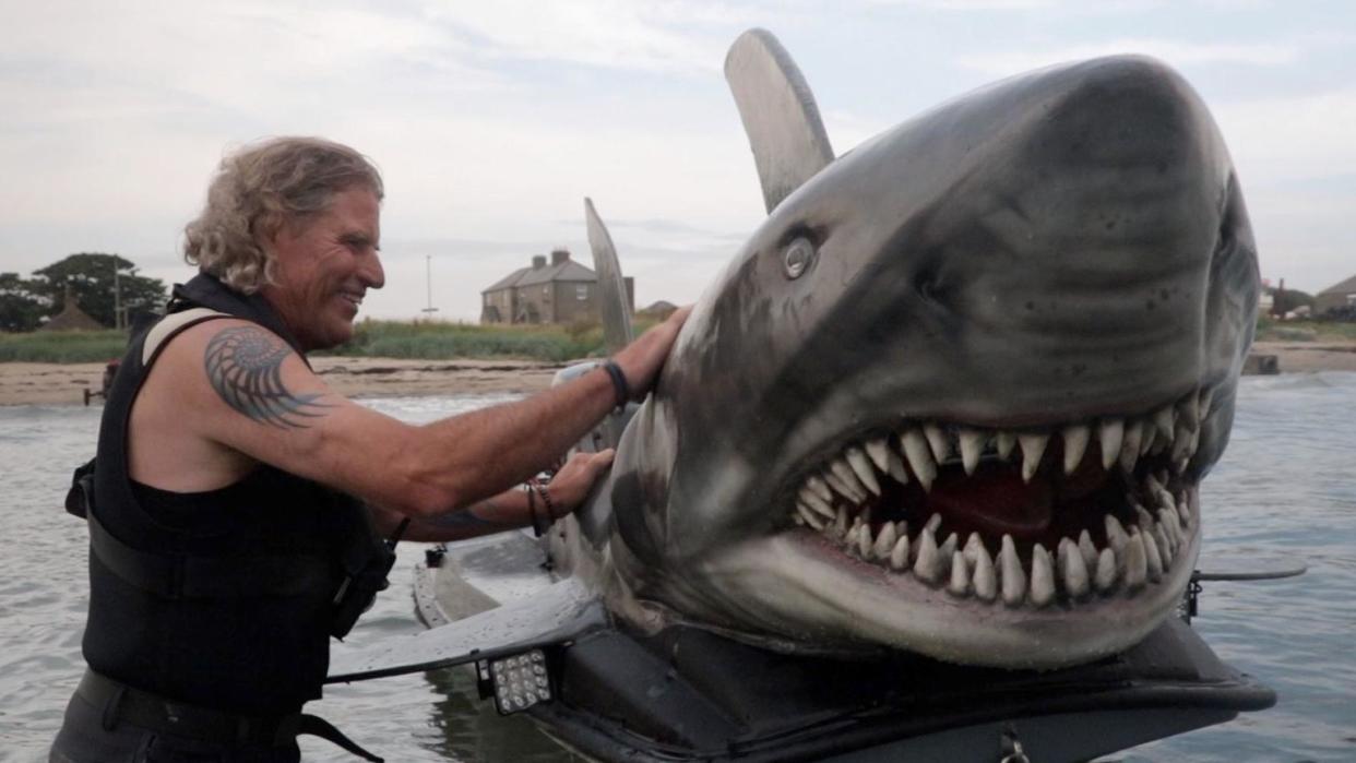 Fin Bowren in the ocean with his shark boat, he stands beside it and pets the head