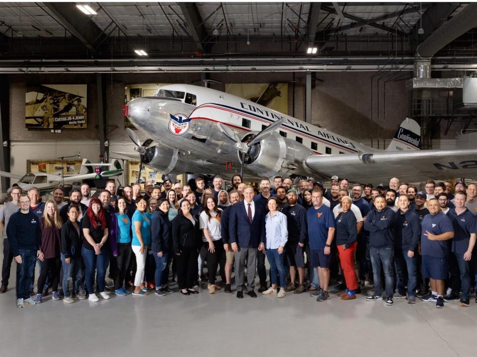 A group of people standing in a hanger in front of large metal plane with two propellers under its wings.