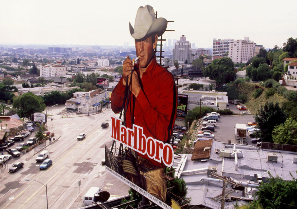 A large billboard featuring the Marlboro Man, a cowboy in a white hat and red shirt, lighting a cigarette with a cityscape in the background