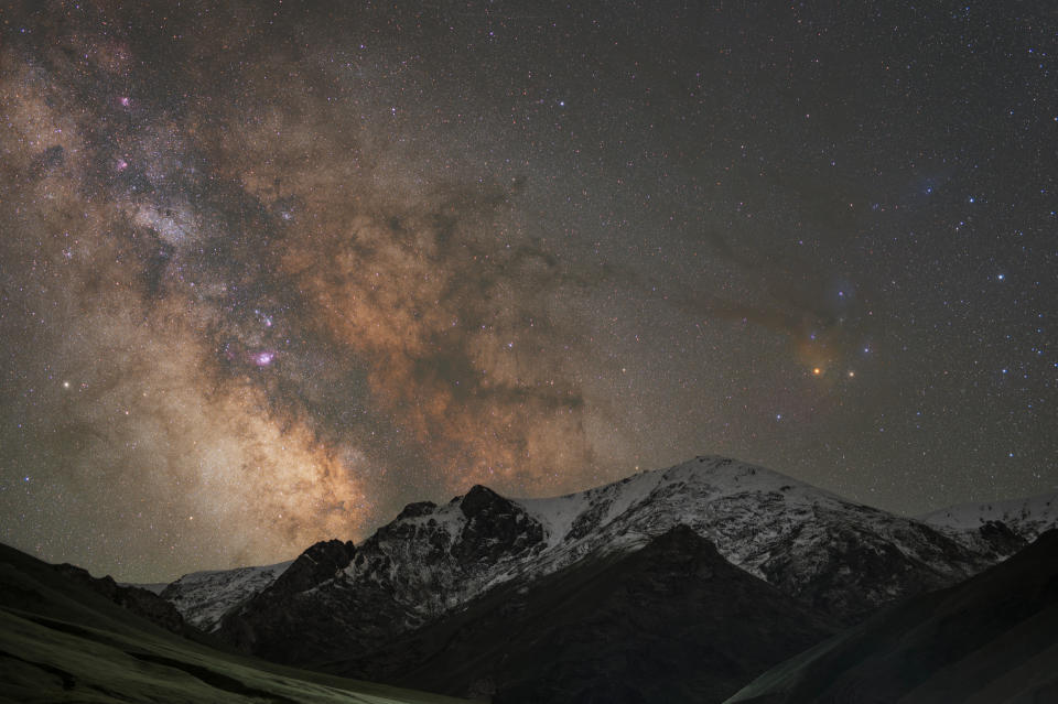 At the bottom of the image are tall, snow-capped mountains and above the Milky Way rises densely with stars up in the sky.